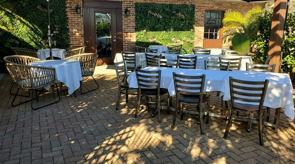 Outdoor restaurant patio with white-clothed tables and chairs on a brick floor, bathed in sunlight.