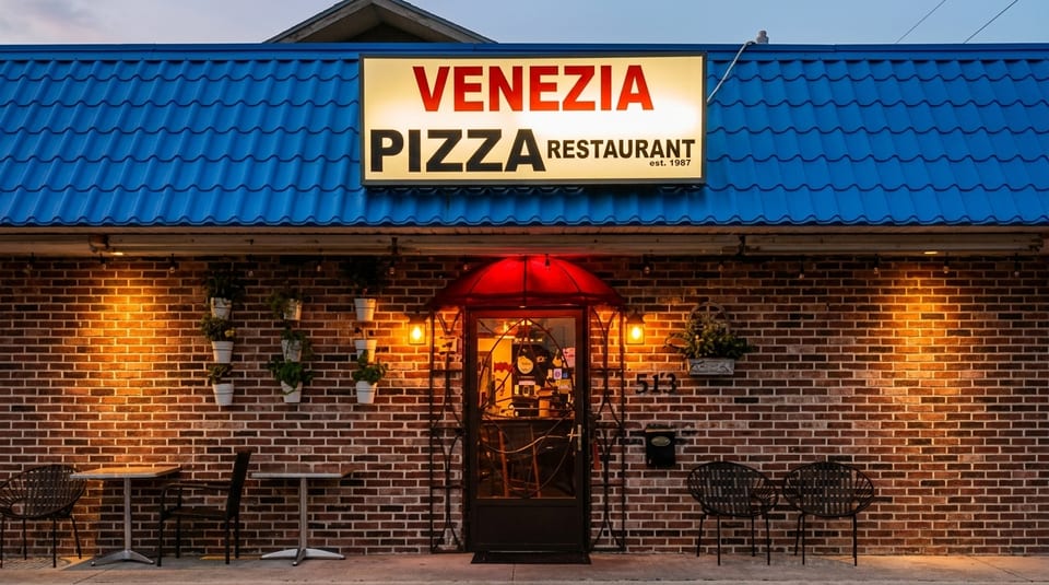 Venezia Pizza Restaurant's illuminated sign above a brick facade with outdoor seating at dusk.